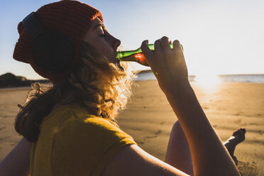 Teenage girl wearing beanie and headphones sitting on the beach, having a drink - UUF08534