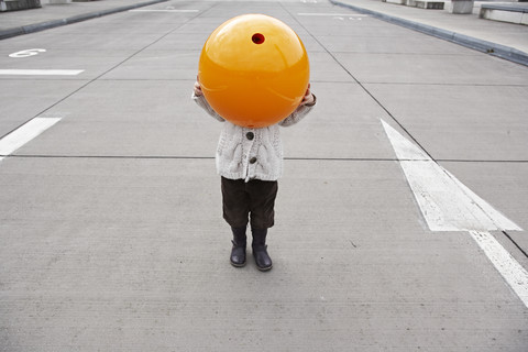 Kleines Mädchen steht auf der Straße und versteckt sich unter einer orangefarbenen Kugel, lizenzfreies Stockfoto