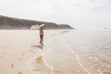 Mature woman walking on the beach lifting a blanket - UUF08563