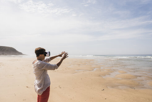 Mature woman standing on the beach wearing VR glasses - UUF08584