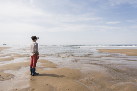 Reife Frau steht am Strand und trägt eine VR-Brille, lizenzfreies Stockfoto