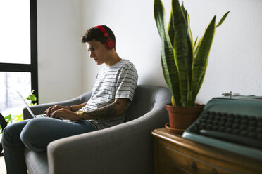 Teenage boy with headphones sitting on couch at home using laptop - EBSF01767