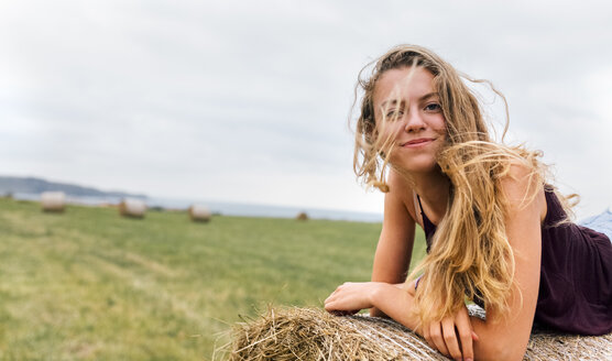 Porträt eines lächelnden blonden Teenagers mit wehenden Haaren auf einem Strohballen liegend - MGOF02437