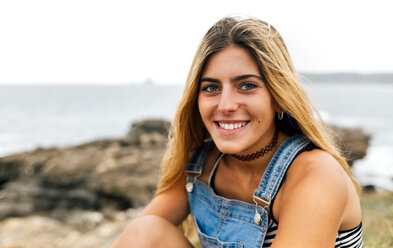 Porträt eines lächelnden Teenagers an der Küste sitzend - MGOF02450