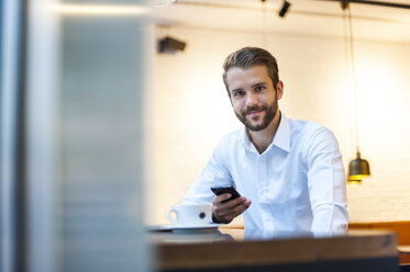 Portrait of smiling businessman with cell phone in a cafe - DIGF01232