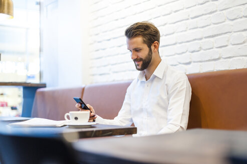 Lächelnder Geschäftsmann mit Blick auf sein Handy in einem Café - DIGF01251