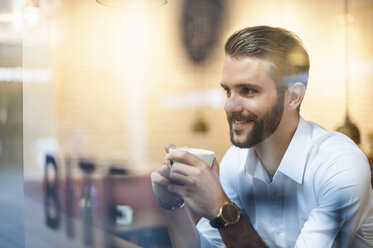 Lächelnder Geschäftsmann hält eine Tasse Kaffee hinter einer Fensterscheibe in einem Café - DIGF01270