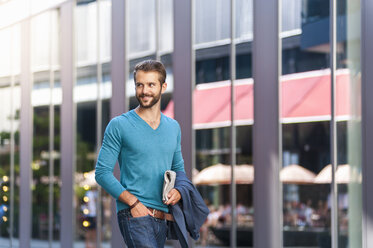 Smiling young man holding newspaper walking in the city - DIGF01303