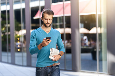 Young man walking with cell phone and newspaper in the city - DIGF01304