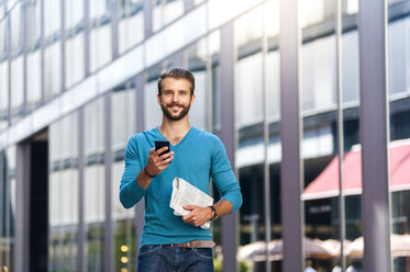 Lächelnder junger Mann mit Mobiltelefon und Zeitung in der Stadt - DIGF01305