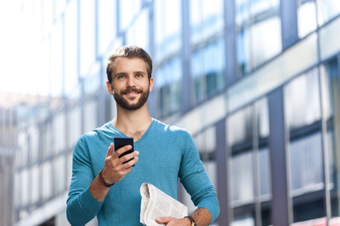 Smiling young man holding cell phone and newspaper in the city stock photo