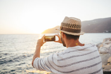 Griechenland, Kykladeninseln, Amorgos, Mann fotografiert den Sonnenuntergang mit einem Smartphone am Meer - GEMF01032