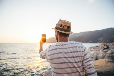 Griechenland, Kykladeninseln, Amorgos, Mann fotografiert den Sonnenuntergang mit einem Smartphone am Meer, lizenzfreies Stockfoto