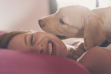 Happy young woman lying on bed playing with her Labrador Retriever puppy - SKCF00203