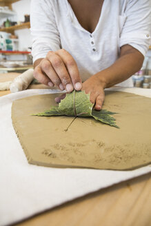 Woman using a leaf to print a draw in terracota in a ceramics workshop - ABZF01245