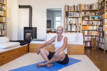 Relaxed woman sitting on gym mat in living room - JUNF00699