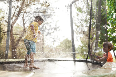 Junge springt auf Trampolin, während sein kleiner Bruder ihn mit Wasser bespritzt - VABF00783