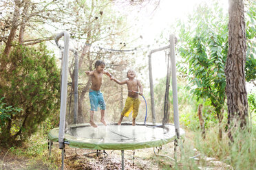 Two little boys jumping on trampoline while splashing with water from garden hose - VABF00788