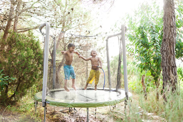 Zwei kleine Jungen springen auf einem Trampolin, während sie mit Wasser aus einem Gartenschlauch bespritzt werden - VABF00788