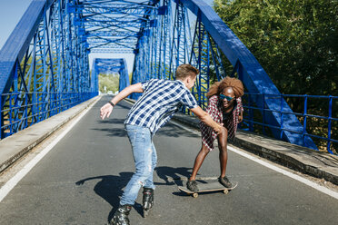 Glückliches junges Paar mit Inline-Skates und Skateboard auf einer Brücke - KIJF00816