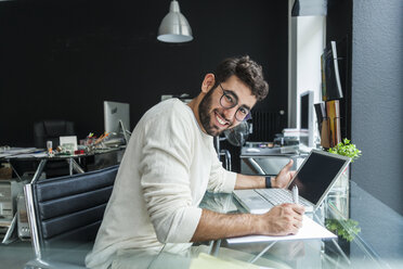 Porträt eines lächelnden jungen Mannes, der an einem Schreibtisch in einem modernen Büro mit einem Laptop arbeitet - TCF05112