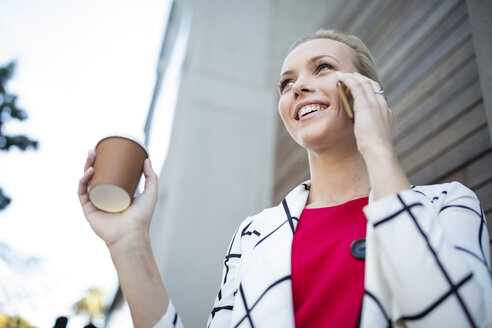 Lächelnde Frau mit Kaffee zum Mitnehmen, die mit ihrem Handy telefoniert - ZEF10485