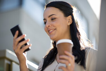 Lächelnde Frau mit Kaffee zum Mitnehmen schaut auf ihr Handy - ZEF10495