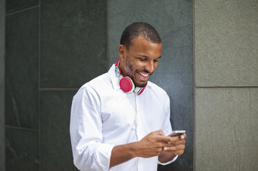 Lächelnder Geschäftsmann mit Kopfhörern, der auf sein Smartphone schaut - DIGF01332