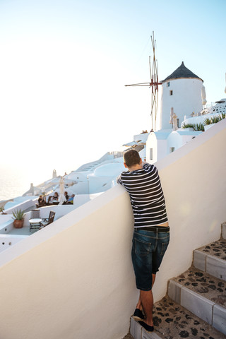 Griechenland, Santorini, Oia, Rückenansicht eines Mannes, der an einer Mauer lehnt und den Sonnenuntergang genießt, lizenzfreies Stockfoto