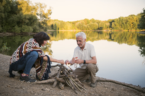 Älteres Paar zündet abends ein Lagerfeuer an einem See an, lizenzfreies Stockfoto