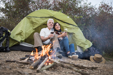 Happy senior couple sitting at campfire with wine at a tent - ONF01049