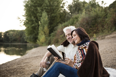 Glückliches Seniorenpaar mit Wein und Buch am See, lizenzfreies Stockfoto