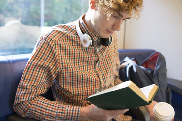 Young man with coffee to go sitting in a train reading a book - FMOF00127