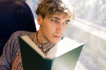 Nachdenklicher junger Mann mit Buch, der in einem Zug sitzt und durch das Fenster schaut - FMOF00137