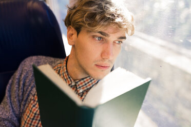 Pensive young man with book sitting in a train looking through window - FMOF00137