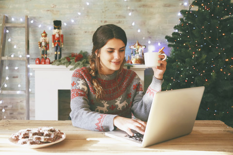 Lächelnde Frau mit einer Tasse Kaffee am Laptop zur Weihnachtszeit, lizenzfreies Stockfoto