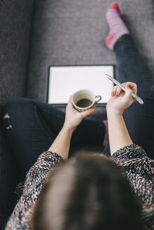 Draufsicht auf eine junge Frau, die mit einem Tablet und einer Tasse Kaffee auf der Couch sitzt und einen Stift hält - LCUF00049