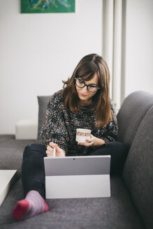 Young woman sitting on the couch with cup of coffee drawing on her tablet - LCUF00050