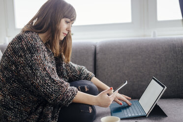 Junge Frau sitzt auf der Couch und benutzt Tablet und Smartphone - LCUF00055