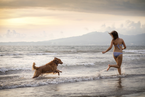 Mexiko, Nayarit, Junge Frau im Bikini spielt mit ihrem Golden Retriever Hund am Strand, lizenzfreies Stockfoto