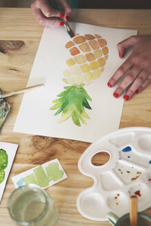 Eine Frau malt mit der Hand ein Aquarell einer Ananas am Schreibtisch in ihrem Atelier - RTBF00438