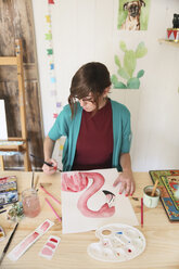 Frau malt ein Aquarell eines Flamingos auf dem Schreibtisch in ihrem Atelier - RTBF00450