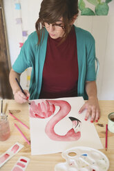 Frau malt ein Aquarell eines Flamingos auf dem Schreibtisch in ihrem Atelier - RTBF00451