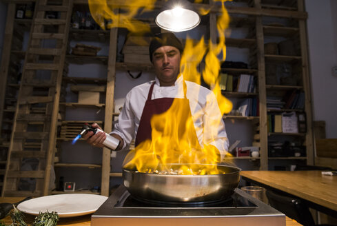 Mann flambiert Rinderbäckchen mit sautiertem Gemüse in einer Pfanne mit einer Küchenfackel - ABZF01362