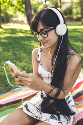 Junge Frau sitzt auf einer Decke im Park und hört Musik - DAPF00370
