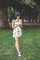 Young woman with headphones walking in park looking at cell phone - DAPF00392