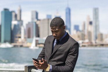 USA, Brooklyn, Geschäftsmann schaut auf sein Handy vor der Skyline von Manhattan - GIOF01478
