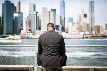 USA, Brooklyn, back view of businessman sitting on bench in front of Manhattan skyline - GIOF01482