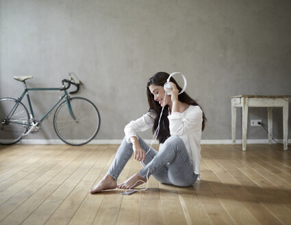 Lächelnde Frau, die zu Hause auf dem Holzboden sitzt und mit Kopfhörern Musik hört - FMKF03098