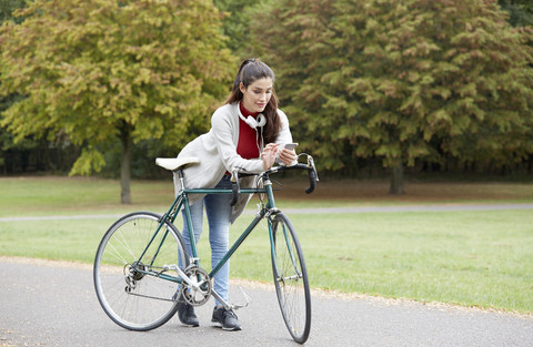 Frau mit Fahrrad in einem herbstlichen Park schaut auf ihr Handy, lizenzfreies Stockfoto